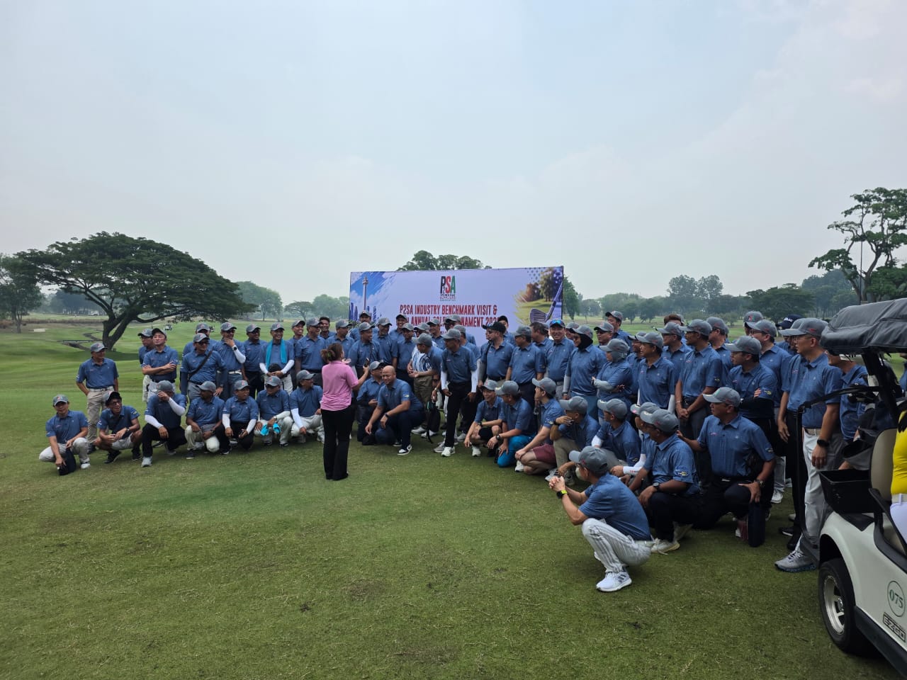 A large group of golfers in matching blue shirts and grey caps gathered on a fairway in front of the tournament banner.