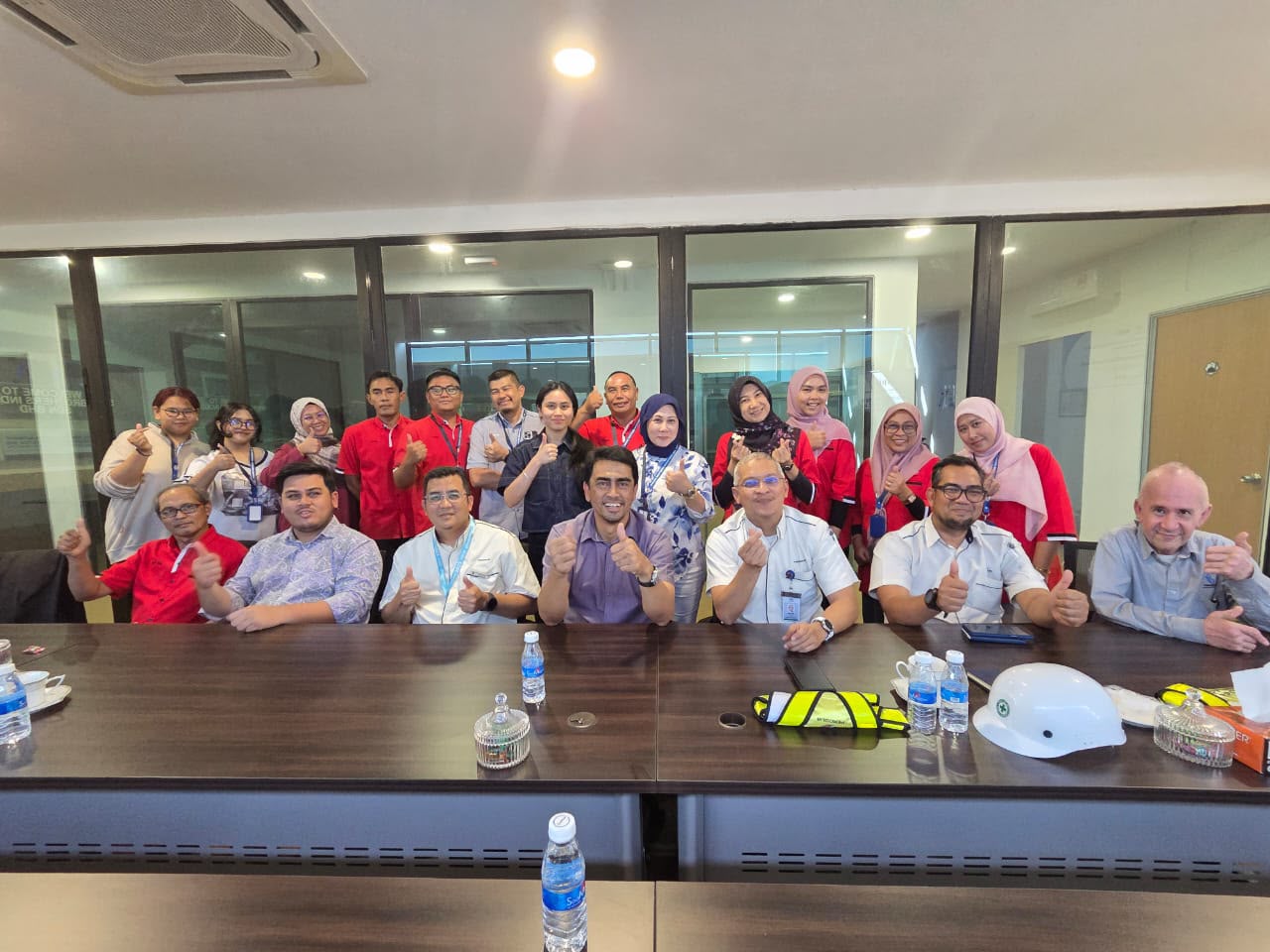 Perodua's visitors and SBI staff in red shirts giving a thumbs-up around a meeting table with a safety helmet and high-visibility vest on it.